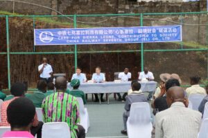 Outdoor community event with five panelists seated at a long table under a blue banner, addressing a seated audience behind a green fence.
