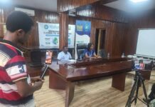ICPNC, UNDP Join Forces to Strengthen Conflict Prevention and National Cohesion in Sierra Leone Man in a striped shirt filming a panel discussion in a wood-paneled conference room, two people seated at a long table with banners behind them.