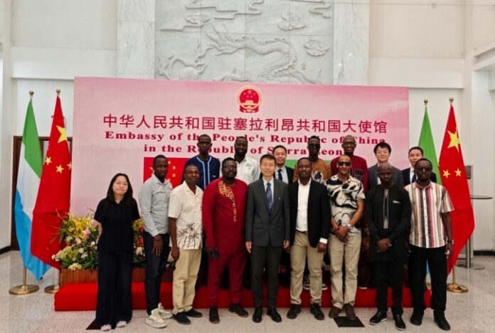 Group of people posing for a photo in front of the Chinese Embassy banner, flanked by flags, on a red carpet.