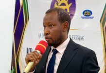 NASSIT DG Calls for Stronger Media Partnership to Improve Social Security Awareness and Compliance Man in a dark suit and blue tie speaks into a red microphone at a formal event, with a banner and plants in the background.