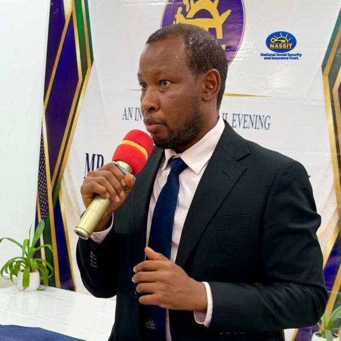 Man in a dark suit and blue tie speaks into a red microphone at a formal event, with a banner and plants in the background.
