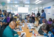 NPHA, IMC Train Journalists on Responsible Reporting as Sierra Leone Strengthens Ebola Preparedness A crowded conference room with many people seated around a long table, cameras filming a presentation on a large screen at the front.