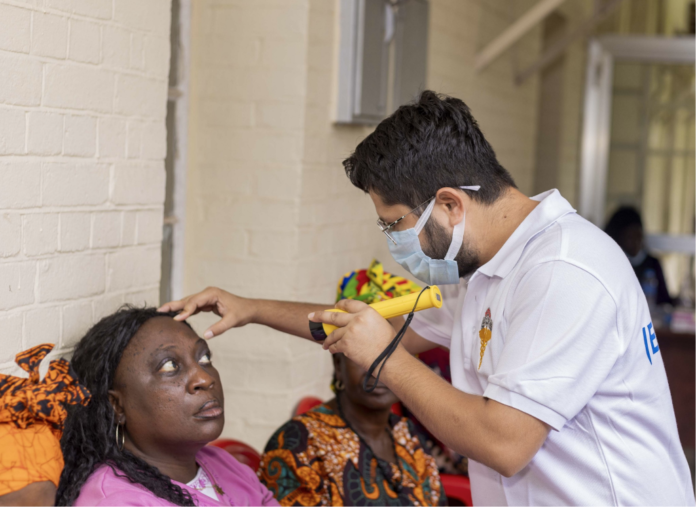 Clinician in a white polo and mask uses a handheld diagnostic device on a woman's forehead during a health screening.