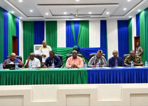 Panel of men seated at a long table during a formal meeting, with green and blue drapes in a decorated hall and water bottles on the table.