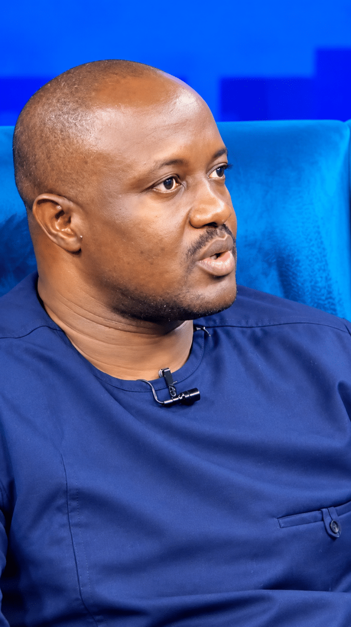Man wearing a navy blue shirt, speaking with a lavalier mic attached to his collar, seated against a blue backdrop.