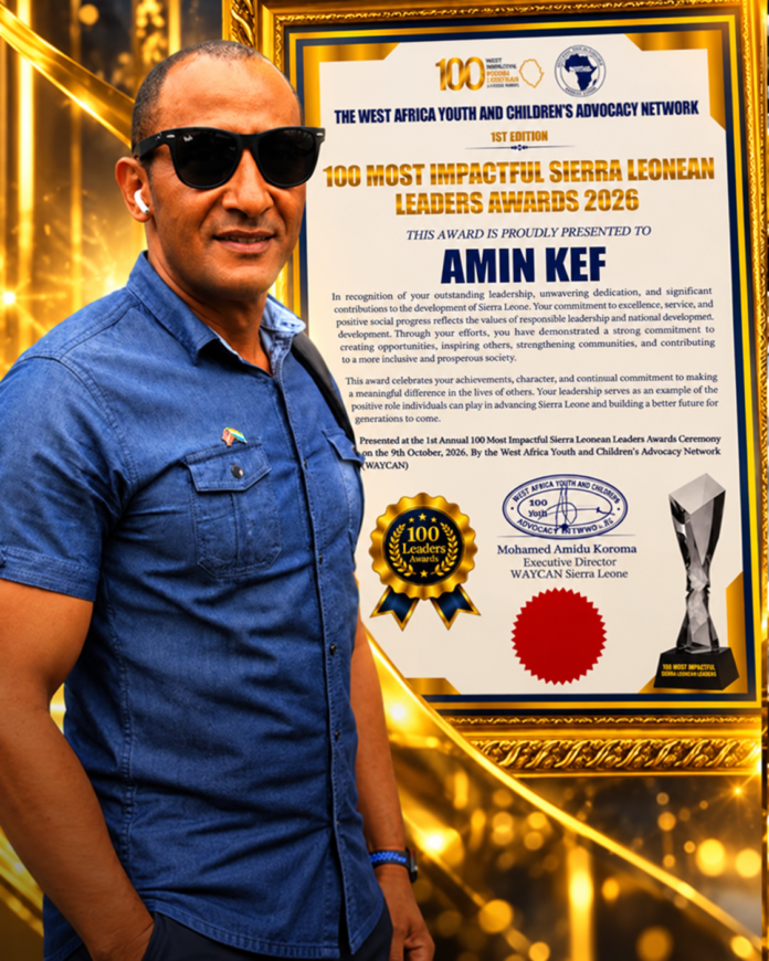 Man wearing sunglasses and a blue shirt poses next to a large gold-framed award certificate for the '100 Most Impactful Sierra Leonean Leaders Awards 2026'. It features a badge and text including the name 'AMIN KEF'.