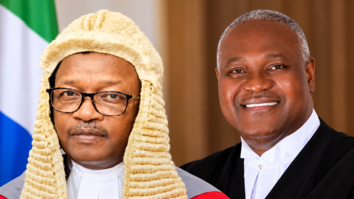 Two Black men in formal judicial attire posing for a portrait; one wears a cream legal wig and glasses.
