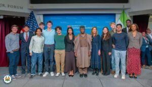 Group of diverse young adults posing for a group photo on a stage with flags and a Harvard Institute of Politics backdrop in the background while smiling for the camera.