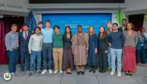 Group of diverse young adults posing for a group photo on a stage with flags and a Harvard Institute of Politics backdrop in the background while smiling for the camera.