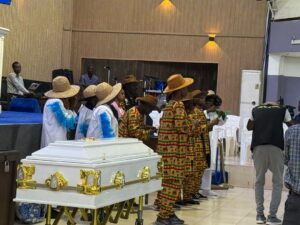 White ceremonial casket with gold accents at a funeral, surrounded by mourners in colorful traditional clothing and wide-brim hats inside a hall.