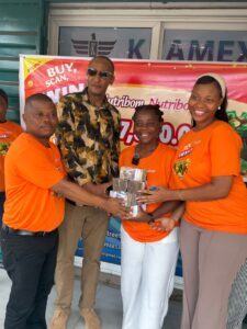 Group of four people outside a shop, two women in orange shirts hand a stack of cash to a smiling recipient in an orange shirt, with a man in sunglasses nearby and another man in orange on the left side.