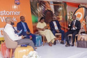 Five panelists seated on a stage during a corporate discussion under an orange event backdrop with branding in the background, microphones within reach, and water bottles on the floor nearby.] ,