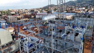 Industrial construction site with steel framework, scaffolding, and a large cylindrical tank; city buildings and hills in the background.