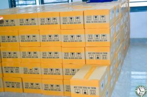 Piled orange shipping boxes in a clean warehouse, each box labeled with handling icons (upright arrows, umbrella for keep dry, fragile) and dimensions.