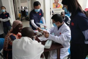 Medical team wearing masks consults with patients at a clinic table, reviewing papers and taking notes, outdoors in a health outreach setting.