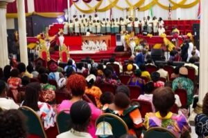 Audience watching a choir perform on a decorated stage with red carpet and gold fabric drapes.