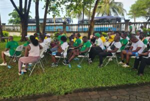 | The Calabash Newspaper People seated on white folding chairs outdoors under trees at an event; many wear green shirts, with buses visible in the background.