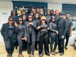 Group of about 16 people dressed in black, posing on steps outside a storefront for a group photo, many wearing sunglasses.