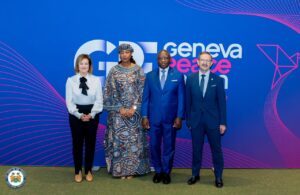 | The Calabash Newspaper Four professionals in formal attire pose in front of a blue Geneva Peace event backdrop with pink lines and logo.",
