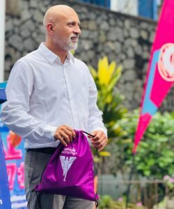 | The Calabash Newspaper Man in a white shirt holds a purple drawstring bag at an outdoor marathon event onlookers in the background.