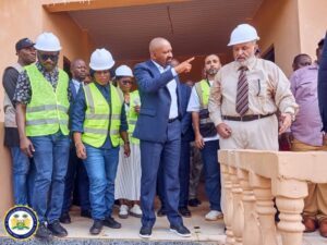 | The Calabash Newspaper Group of construction workers and officials on a building site, wearing hard hats and hi-vis vests; a man in a blue suit points as others listen on a partially built porch.