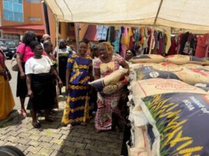 Women in bright dresses chat near stacked sacks of grain at an outdoor market under a tent tallied with clothing stalls in the background.