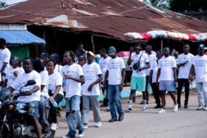 A group of people in white T-shirts walk in a line along a street, with market stalls and a rusted roof behind them in a sunny parade scene.