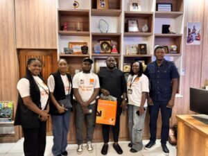 Six people pose for a group photo in an office lobby with wooden display shelves of awards behind them; the man in the center holds an orange gift bag.