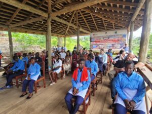 Group of students and adults seated on wooden chairs in a shaded open-air pavilion, attending a hospitality training event with a banner in the background.
