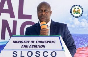 Man in a navy suit speaks into an orange microphone at a podium labeled 'Ministry of Transport and Aviation' with a government seal in the background.