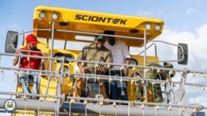 | The Calabash Newspaper Four men stand on a yellow elevated work platform labeled SCIONTEK, inspecting machinery against a blue sky.