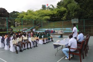 Students in uniform sit on white chairs on an outdoor basketball court, listening to a man speaking at a table with officials nearby.