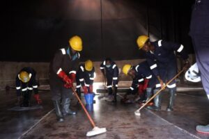 | The Calabash Newspaper Group of workers in yellow hard hats sweeping a wet factory floor at night, wearing reflective gear.