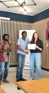 Two people shake hands while a woman receives a certificate at a small indoor awards ceremony, a man claps in the background.