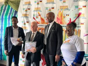 Four people pose at an event with a colorful banner behind them; two men in suits hold wrapped items, a taller man in a suit stands with them, and a woman in a white event shirt smiles nearby.