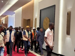 | The Calabash Newspaper Group of people walking through a modern art gallery corridor; a man in a light pink shirt leads, with framed artwork on the walls.