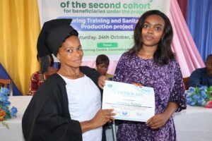 Two women at a certificate ceremony: an older graduate in a black gown and cap on the left, a younger recipient in a purple patterned dress holding a certificate, with a banner in the background.