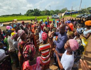 Large group of Africans in colorful traditional clothing dancing and clapping outdoors, with a rural backdrop and tents in the distance.