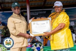 Two men exchange a framed certificate at an outdoor ceremony, one in a beige uniform and green cap, the other in a yellow shirt and white cap.