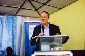 UNDP Deputy Resident Representative delivering remarks at the Orientation Launch Ceremony | The Calabash Newspaper Man in a suit speaks at a podium with a microphone during a formal event. Behind him are banners and a blue drape.