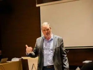 Man in a gray blazer giving a talk in a conference room, gesturing with his right hand.