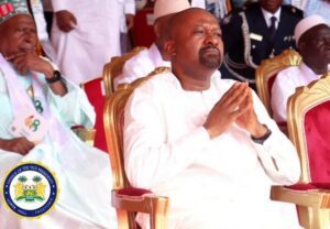 Man in white traditional attire seated with hands pressed together in prayer at a formal ceremony; gold chairs and other attendees in the background.