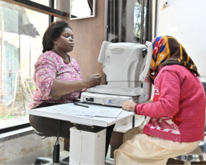 Two women in a clinic: a staff member operating an eye exam device while a patient wearing a colorful headscarf sits beside it for a vision test.