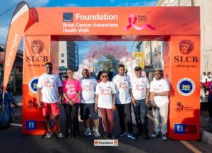 Group of eight volunteers posing under an orange Foundation arch for a Breast Cancer Awareness Health Walk on a sunny street.