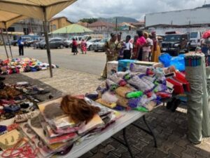 | The Calabash Newspaper Outdoor market scene with stalls under tents selling colorful fabrics and clothing; shoppers mingle near parked cars in a sunny open space.