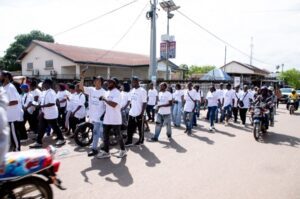 | The Calabash Newspaper A large group of people wearing white T-shirts marching down a sunlit street in a town, with motorcycles and houses in the background.