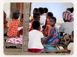 | The Calabash Newspaper Group of young children seated on wooden benches under a shelter, wearing colorful clothes and looking around.