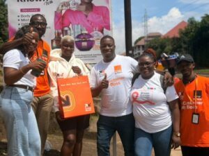 Group of six people outdoors posing for a photo, giving thumbs up; some wear orange uniforms and one wears a breast cancer awareness shirt with a pink ribbon.
