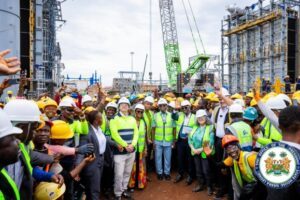 Group of construction workers and officials wearing hard hats and high-visibility vests posing at a busy construction site with cranes and scaffolding in the background.