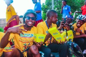Group of smiling youths in yellow shirts at an outdoor festival, holding ONGA Waka Fiesta food boxes and posing for photos.